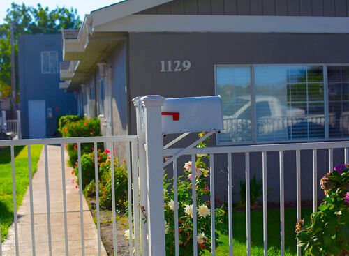 White fence enclosure with mailbox in front of a multiplex building surrounded by green plants